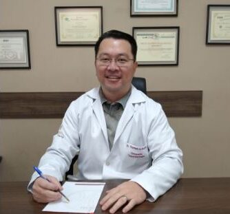 Dr. Thomas Jo Kurita, de jaleco branco, sentado à mesa do consultório, com certificados emoldurados na parede ao fundo.